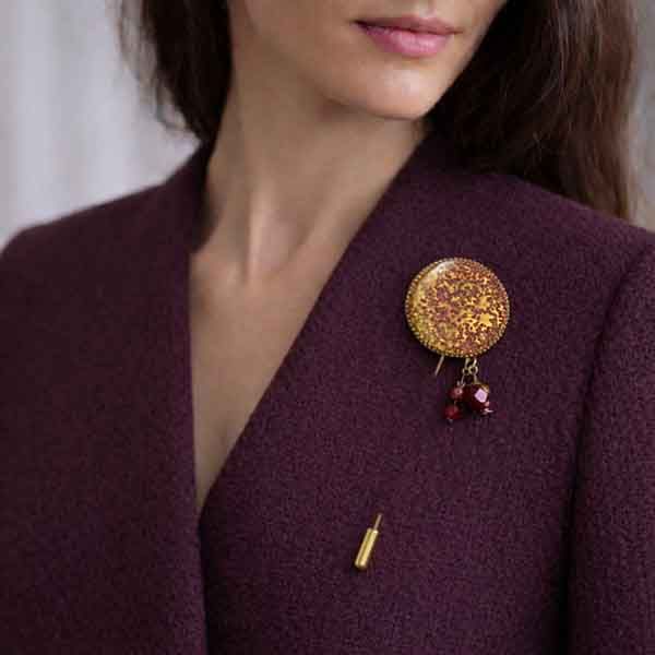 Aguja Planet artesanal con esmalte en tonos dorado y berenjena colocada en chaqueta color berenjena