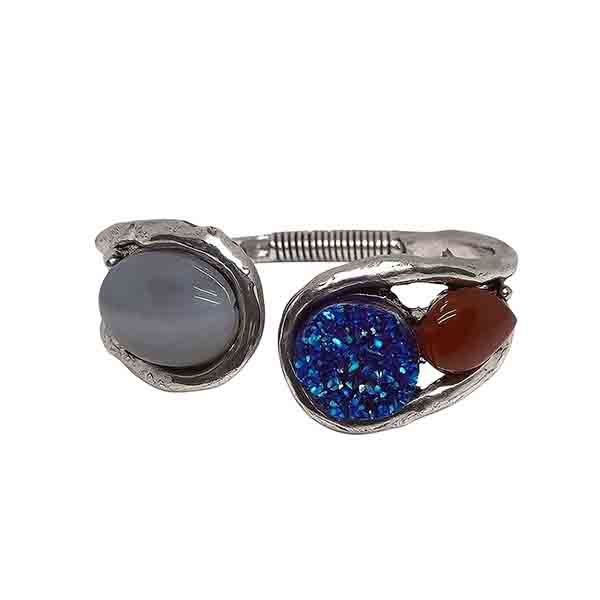 Pulsera Estigia piedras Pulsera Estigia piedras de metal plateado con tres piedras en tonos gris, azul y caramelo sobre fondo blanco.