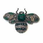 Broche Hayar con cristales verde botella y gris ahumado en forma de abeja sobre fondo blanco, fotografía de producto profesional.