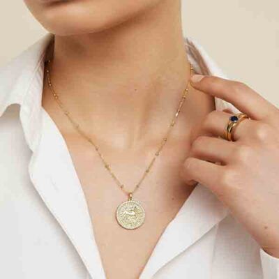 Modelo luciendo el collar horóscopo de Luxenter, una joya dorada con colgante circular grabado, sobre camisa blanca.