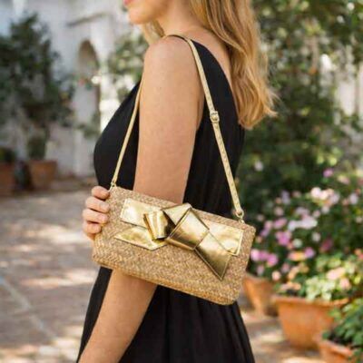 Bolso clutch rafia con lazo dorado y correa larga regulable llevado por modelo con vestido negro