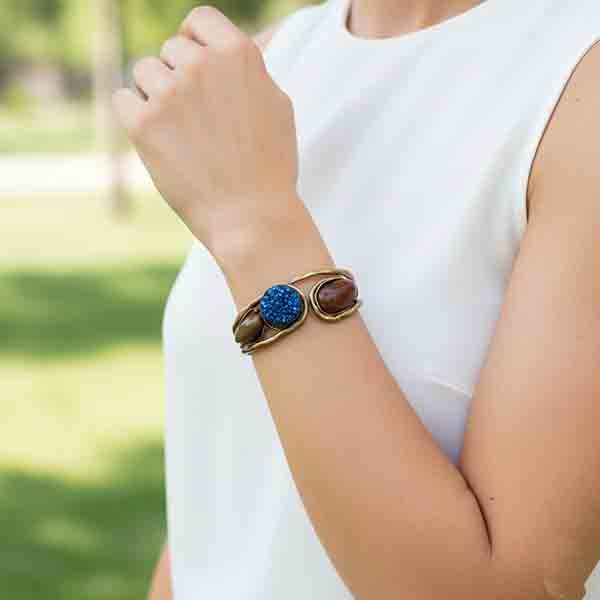 Mujer luciendo la Pulsera Thalassa en metal dorado envejecido con piedras naturales azul y marrón, en un look elegante al aire libre.