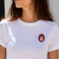 Broche Pamplonica pintado a mano colocado sobre una camiseta blanca. Diseño exclusivo de Turquesa y Más inspirado en San Fermín, Pamplona y la colección Rojo y Blanco de Colores de mi Tierra.